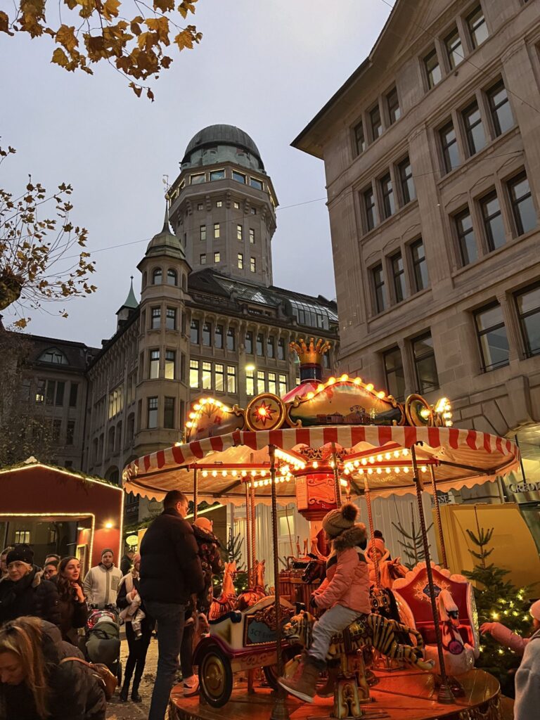 Children riding illuminated vintage carousel at Werdmühleplatz Christmas market in Zurich with Observatory tower in background