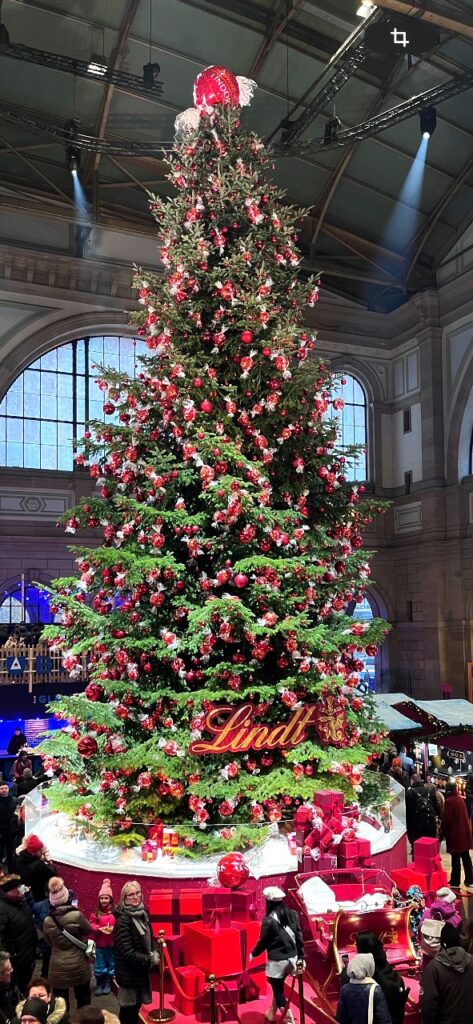 Ten meter tall Lindt chocolate Christmas tree decorated with red ornaments inside Zurich Hauptbahnhof main train station with crowds of visitors and wrapped presents below