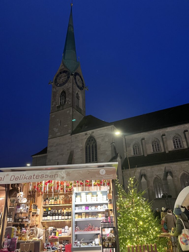 Christmas market wooden stall selling delicatessen and Swiss products with illuminated Fraumünster church tower in background at Münsterhof square, Zurich