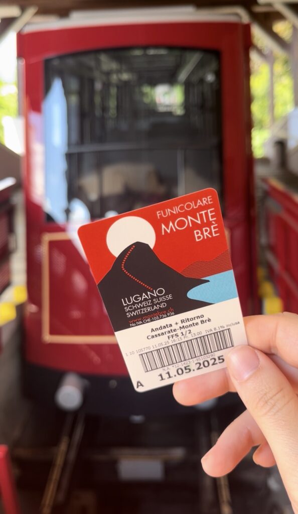 Travel blogger Eleni Kotidou (worldtravellen.com) holding a ticket with the red Monte Bre funicular in the background, Lugano, Switzerland